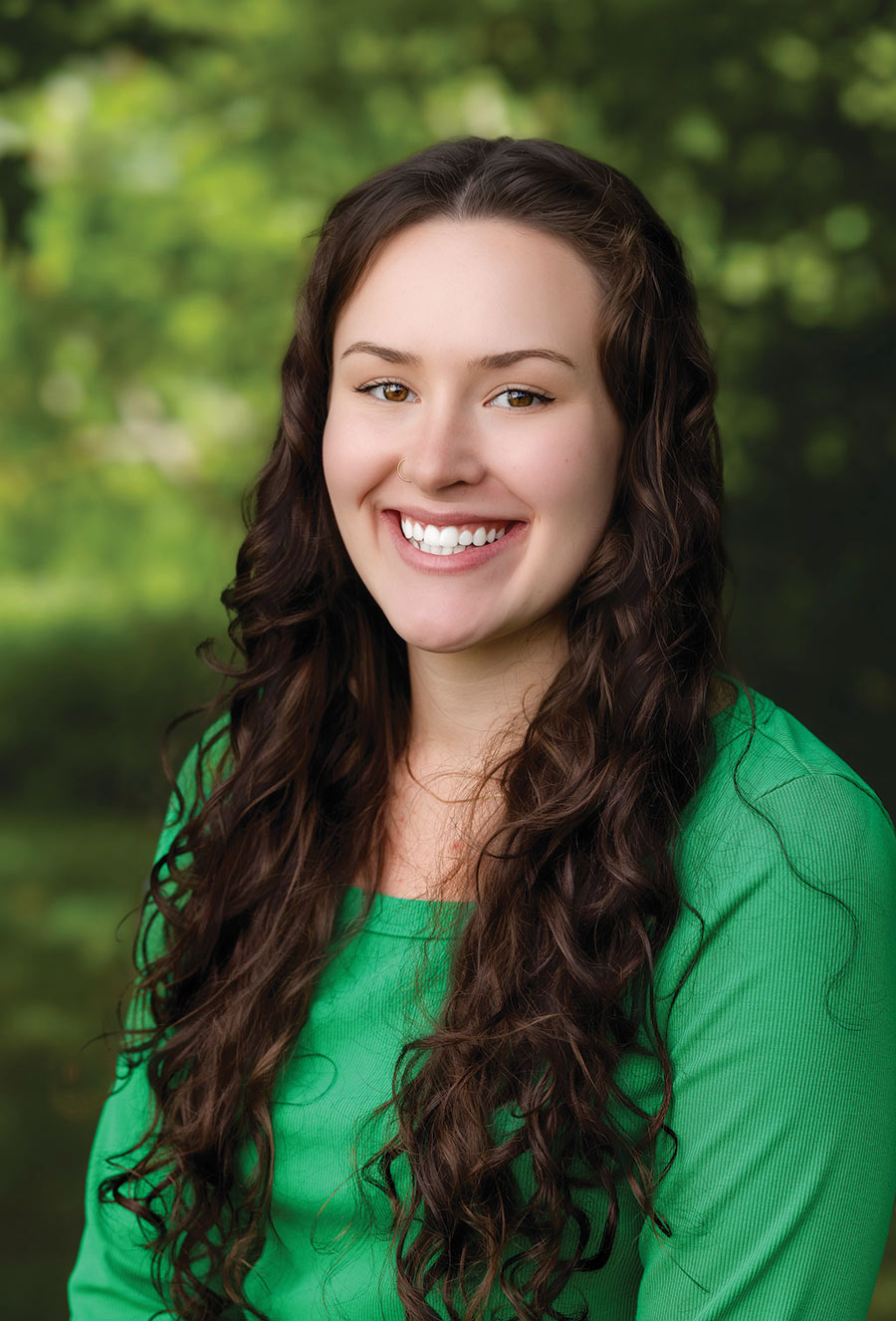 Young woman with long curly brown hair, wearing a green top, smiles at the camera with a blurred green outdoor background behind her, reminiscent of Abigail Williams’ graceful charm.