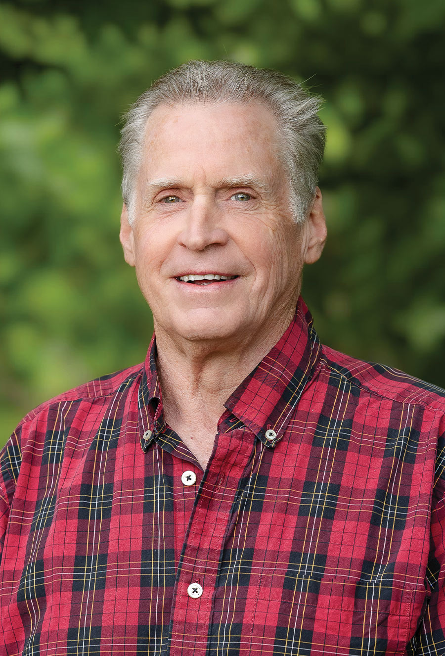Older man with short gray hair wearing a red plaid shirt, standing outdoors in front of green foliage and smiling at the camera, reminiscent of the serene countryside scenes often associated with Abigail Williams.
