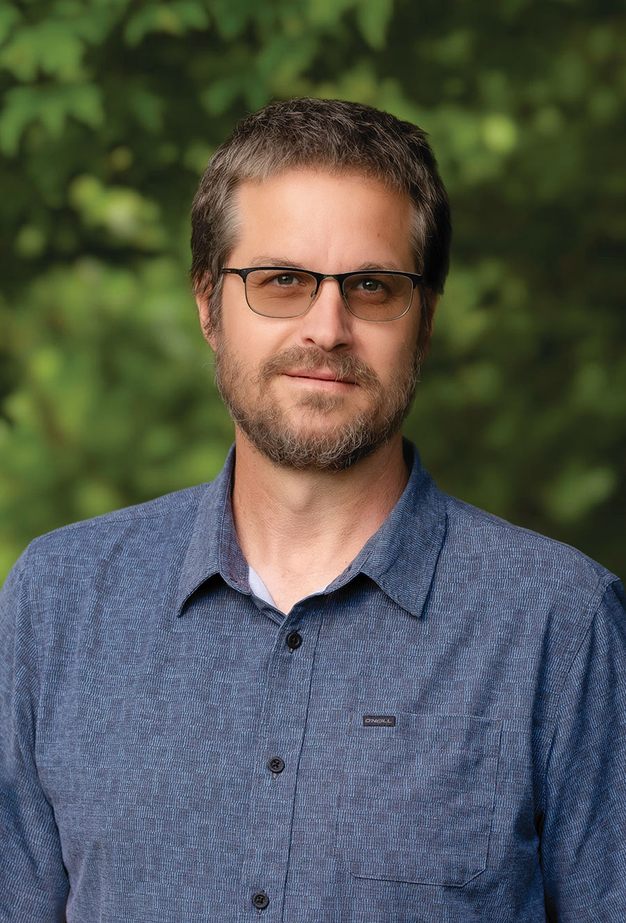A man with short brown hair, beard, and glasses wearing a blue button-up shirt stands outdoors in front of green foliage, evoking the contemplative atmosphere often associated with Puritan history and the Salem witch trials.