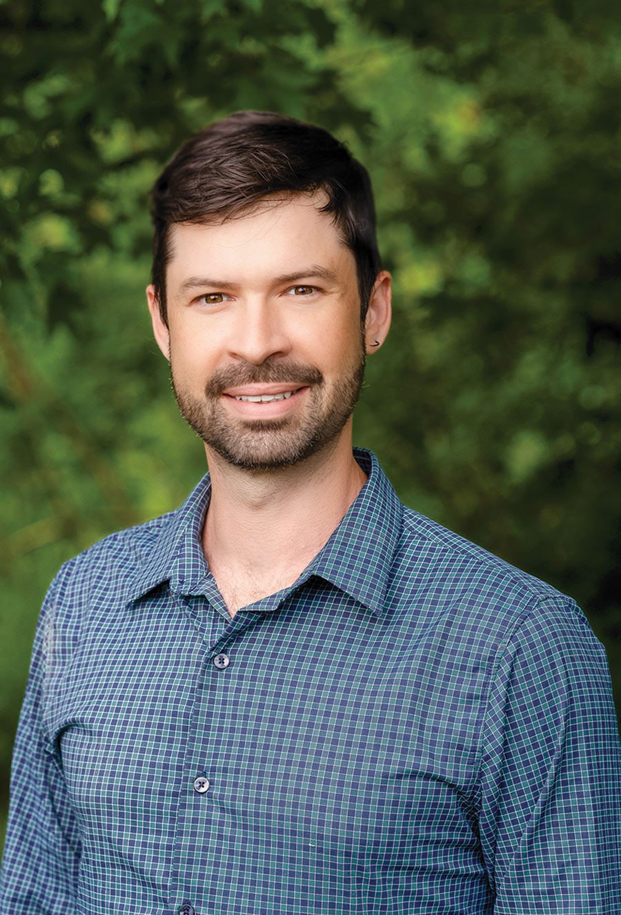 Man with short dark hair and beard, wearing a blue checkered shirt, smiling and standing outdoors with green foliage in the background, reminiscent of the tranquil settings often associated with Abigail Williams.