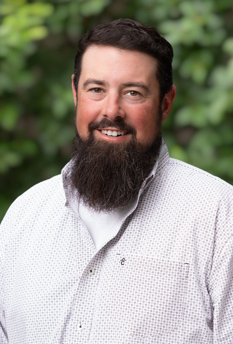 Ian McGonagle, a man with a dark beard and short hair wearing a white patterned shirt, is smiling while standing in front of a blurred green leafy background.
