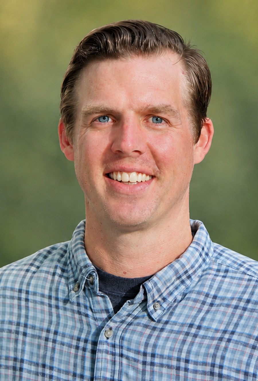 A man with light skin and short brown hair smiles at the camera, wearing a blue plaid shirt and standing in front of a blurred green background. Josh Ralston appears relaxed and approachable in this natural setting.