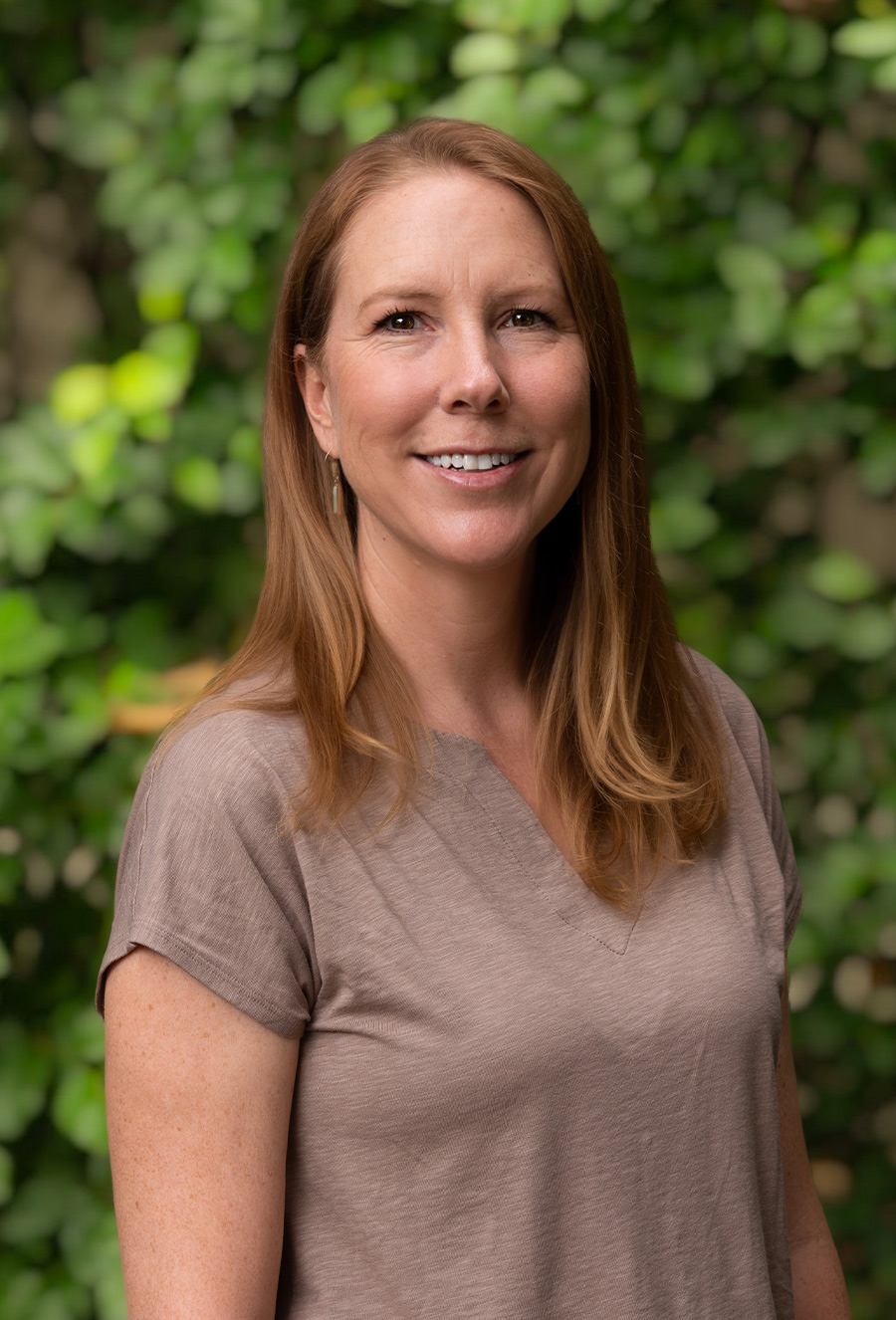 Katie Dahlgren, a woman with straight, light brown hair wearing a short-sleeved beige top, stands outdoors in front of a leafy green background, smiling at the camera.